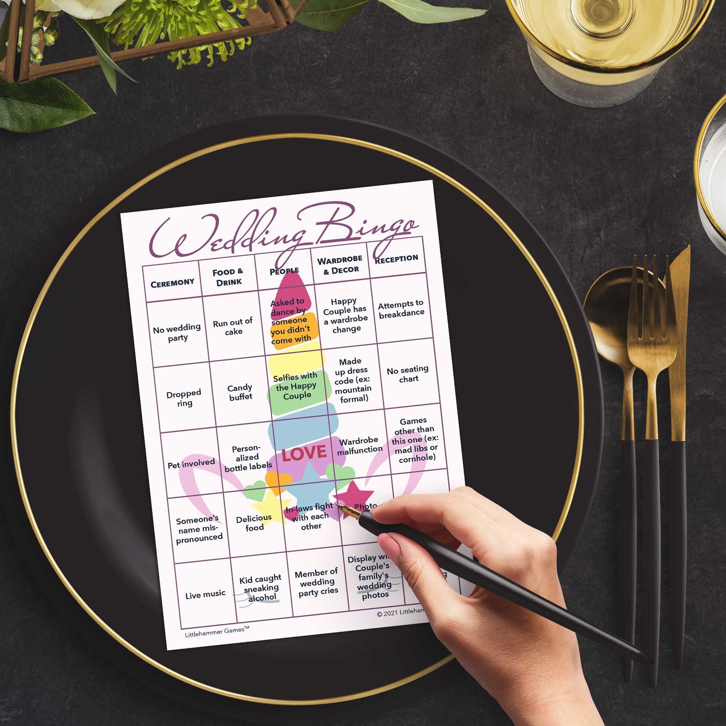 Woman with a pen sitting at a table with a unicorn-themed Wedding Bingo game card on a black and gold plate at a dark place setting