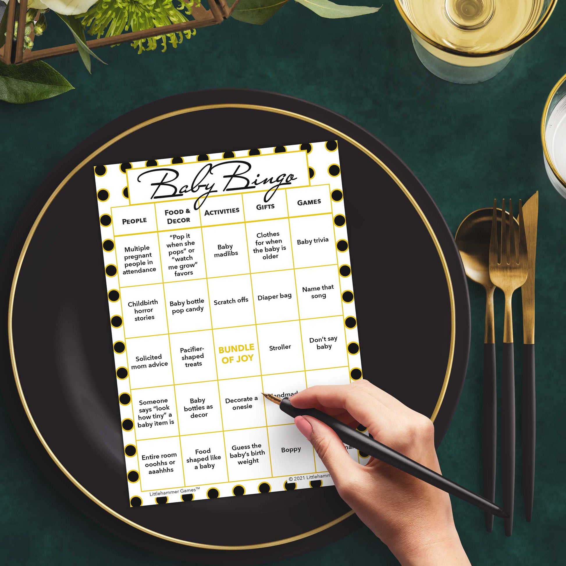Woman with a pen sitting at a table with a black and gold polka dot Baby Bingo game card on a black and gold plate at a dark place setting