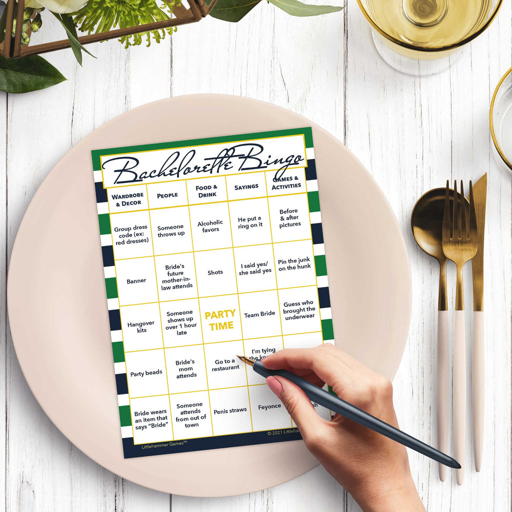 Woman’s hand holding a pen playing a green and navy-striped Bachelorette Bingo game card at a light place setting