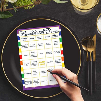 Woman with a pen sitting at a table with a rainbow striped Bachelorette Bingo game card on a gold and black plate on a dark place setting