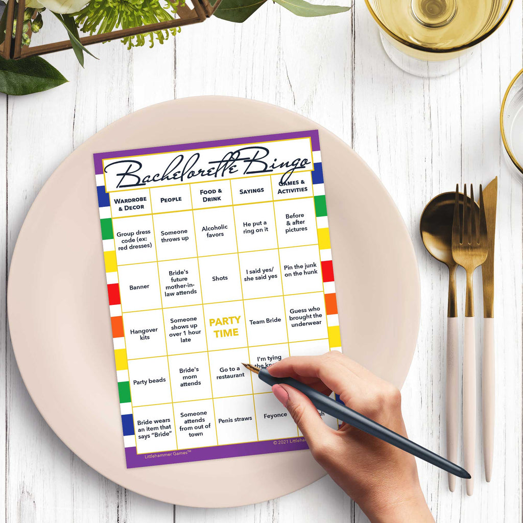 Woman with a pen sitting at a table with a rainbow striped Bachelorette Bingo game card on a gold and black plate on a dark place setting