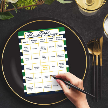 Woman with a pen sitting at a table with a green and navy-striped Bridal Bingo game card on a black and gold plate at a dark place setting