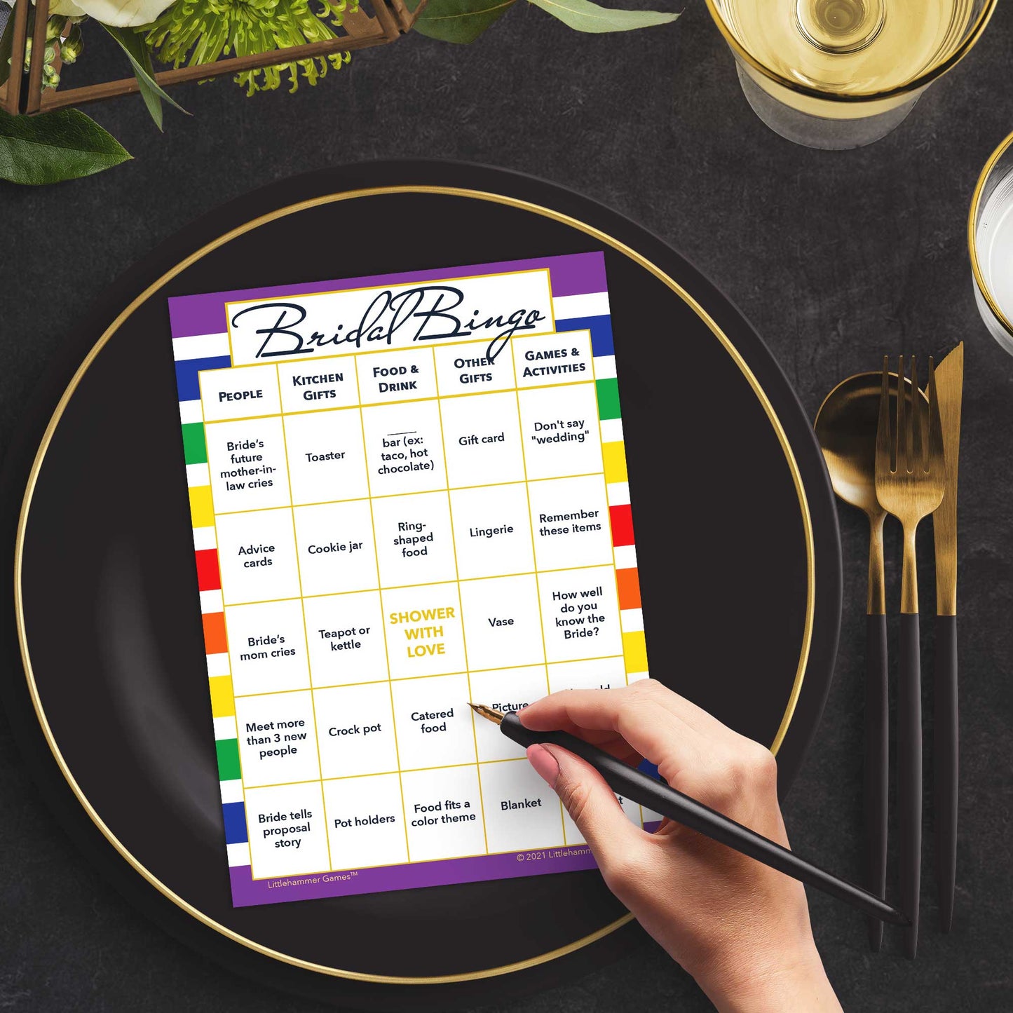 Woman with a pen sitting at a table with a rainbow-striped Bridal Bingo game card on a black and gold plate at a dark place setting